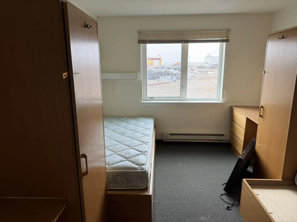A furnished bedroom inside the Frontier camp with a bed, window, desk, and wardrobe